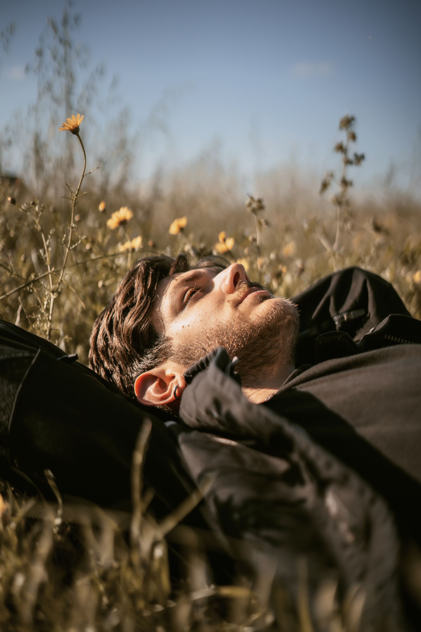 A man with short hair lying down in a meadow with flowers, enjoying a sunny day.